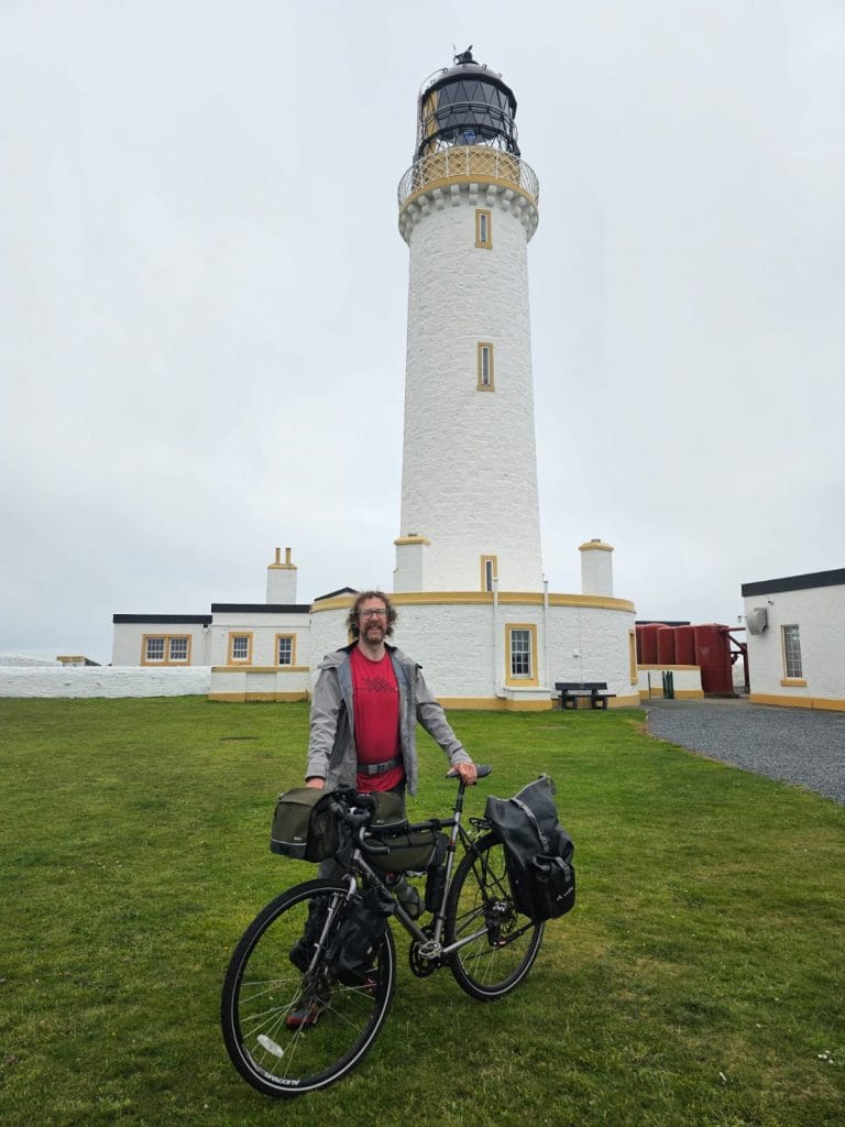 Ally Burns posing in front of a lighthouse with his bike, along his fundraising challenge for Vocal.