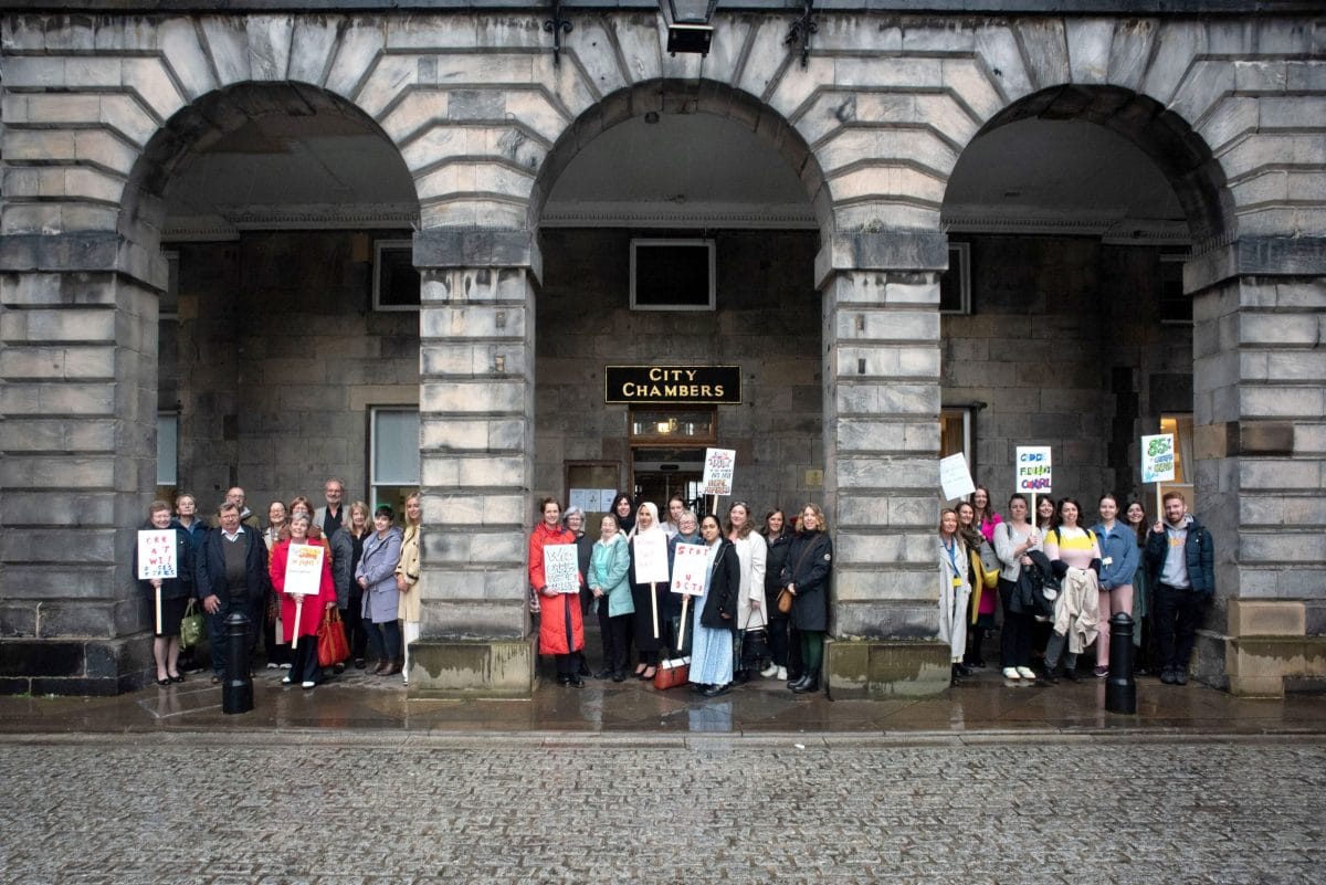 Large group stands under the arches outside the front door of the Edinburgh City Chambers. Some people hold up signs with slogans such as "Who cares about carers?".