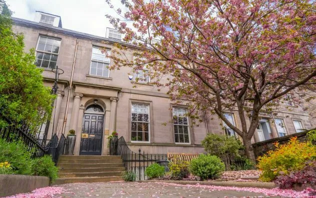 Spring-time view of VOCAL Edinburgh Carers' Hub. It is an old 4-storey building with stairs leading to a black front door. In the front garden is a cherry blossom tree and the pink petals cover the ground.