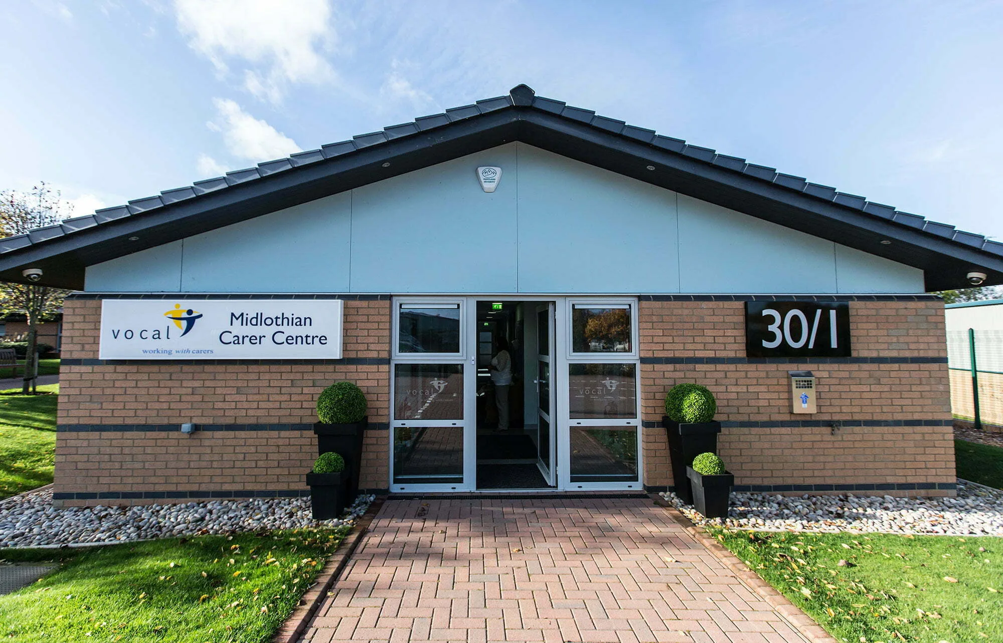 Front view of VOCAL Midlothian Carer Centre. It is a low building with red brick and glass front doors. In front is a stone path and green garden.