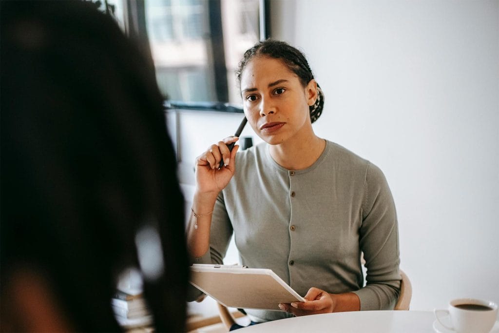 Young woman with a serious look on her face, listening to someone else speak (who cannot be seen). She is holding a notebook and pen.