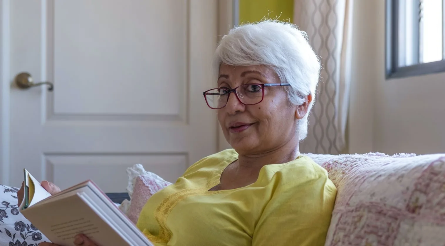 South Asian woman with white hair and glasses sits on the sofa reading a book.