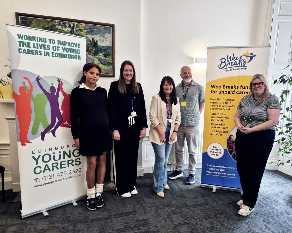 A young carer and two staff members from Vocal, and two from Edinburgh Young Carers, standing in front of banners for their services. They read "working to improve the lives of young carers in Edinburgh" and "Wee Breaks funding for unpaid carers".