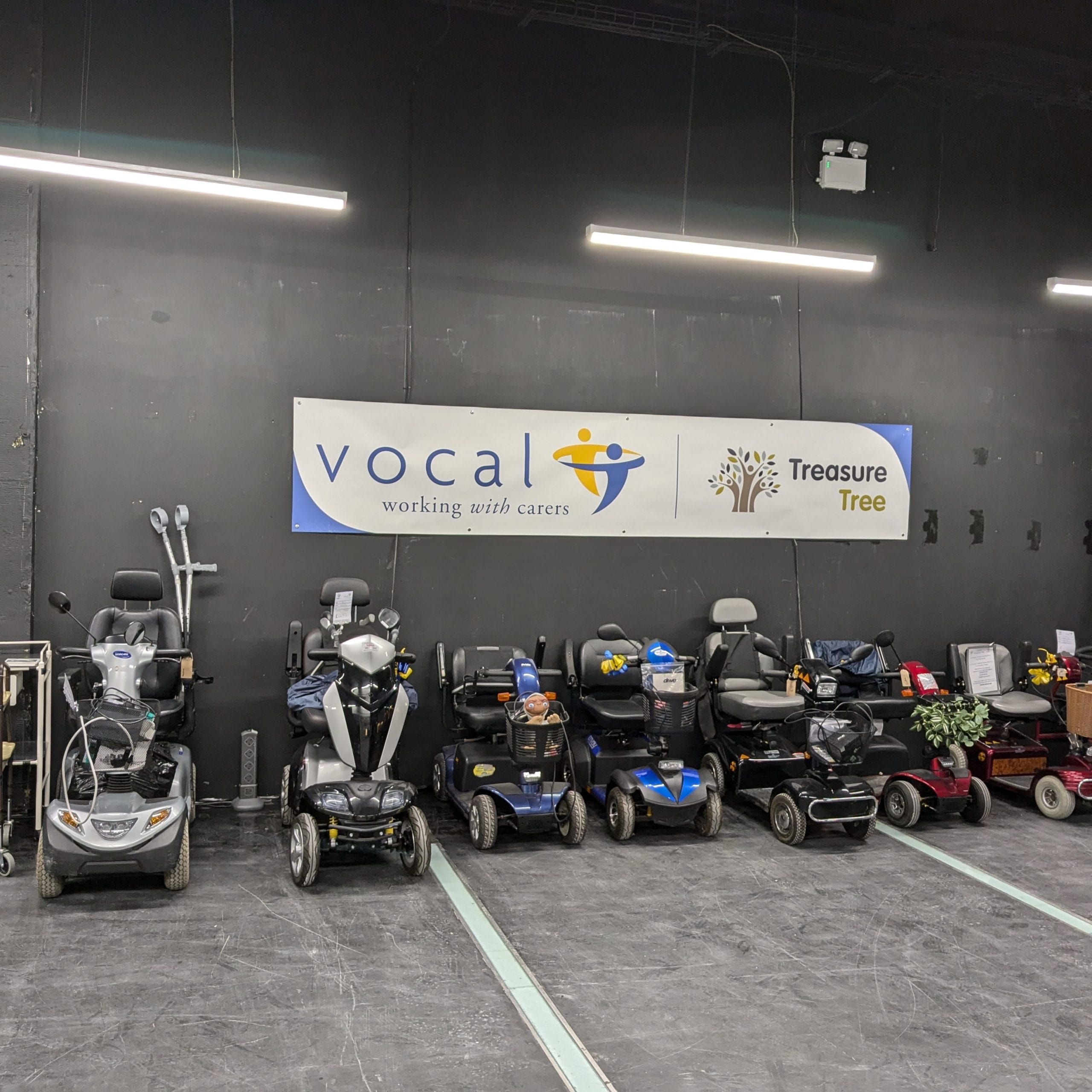 A selection of motorised wheelchairs, scooters and walking aids displayed in Treasure Tree's new unit in Ocean Terminal. The sign on the wall has logos for Vocal and Treasure Tree.